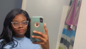 A young Black woman with clear glasses and long dark hair taking a mirror selfie.