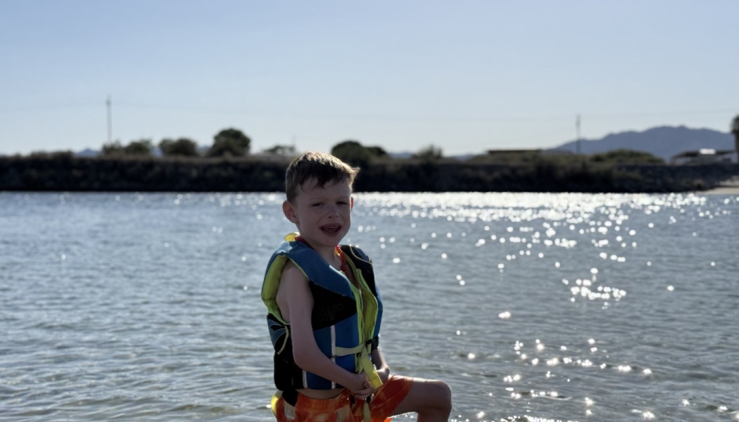 A young boy wearing a life vest smiles while standing in sparkling water on a sunny day.