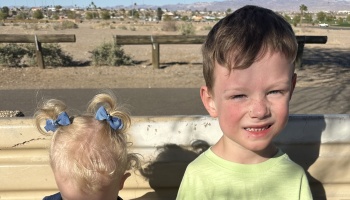 A smiling young boy in a light green shirt sits outdoors next to a toddler with blonde pigtails and blue bows.