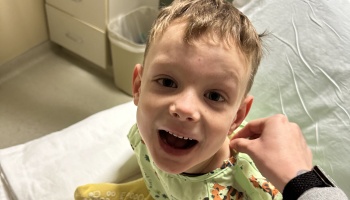 A young boy in a patterned hospital gown smiles brightly while sitting on a hospital bed.