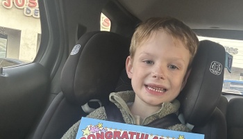A smiling young boy sitting in a car seat holding a colorful "Congratulations" certificate.