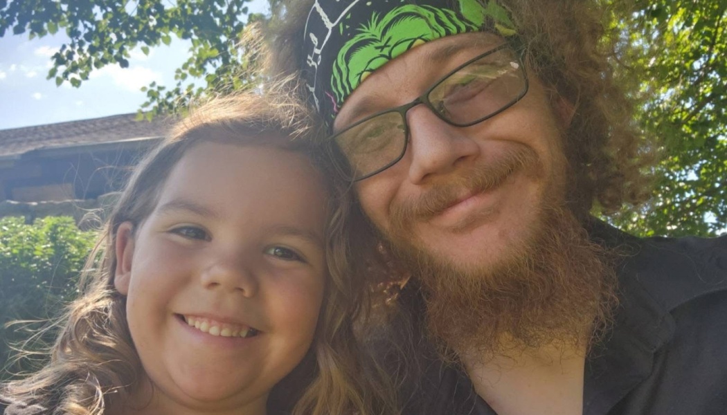A bearded man wearing glasses and a headband smiles next to a young girl outdoors.