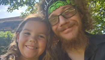 A bearded man wearing glasses and a headband smiles next to a young girl outdoors.