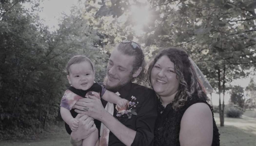 A smiling man and woman holding their cheerful baby outdoors with sunlight filtering through the trees.