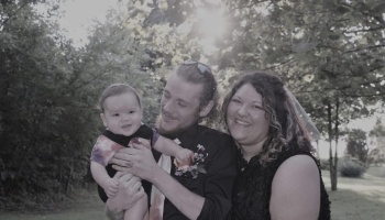 A smiling man and woman holding their cheerful baby outdoors with sunlight filtering through the trees.