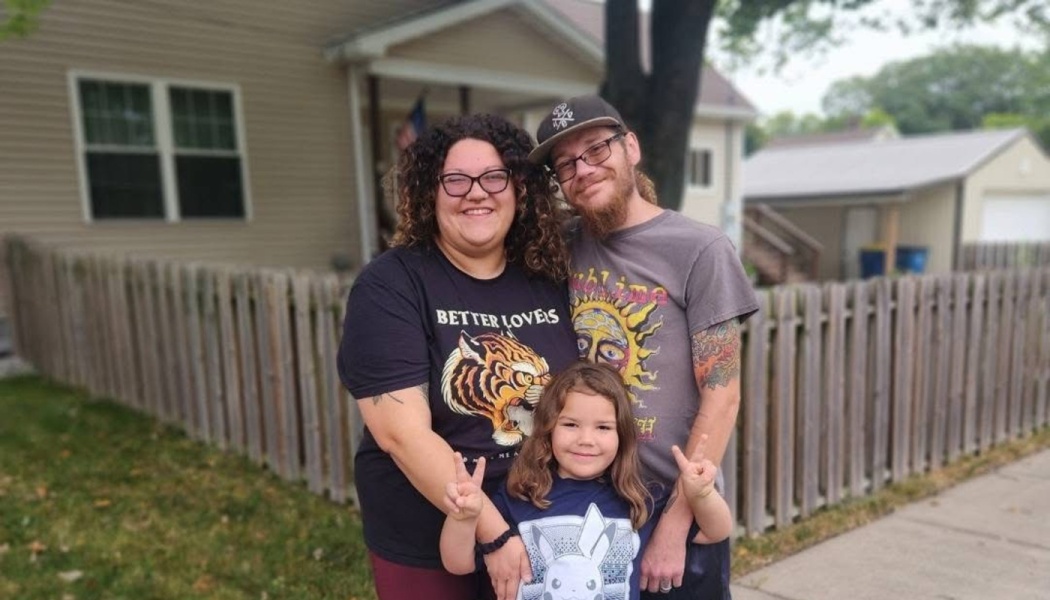 A smiling family of three—a mother, father, and young child making peace signs—poses together outside a house.