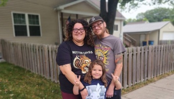 A smiling family of three—a mother, father, and young child making peace signs—poses together outside a house.