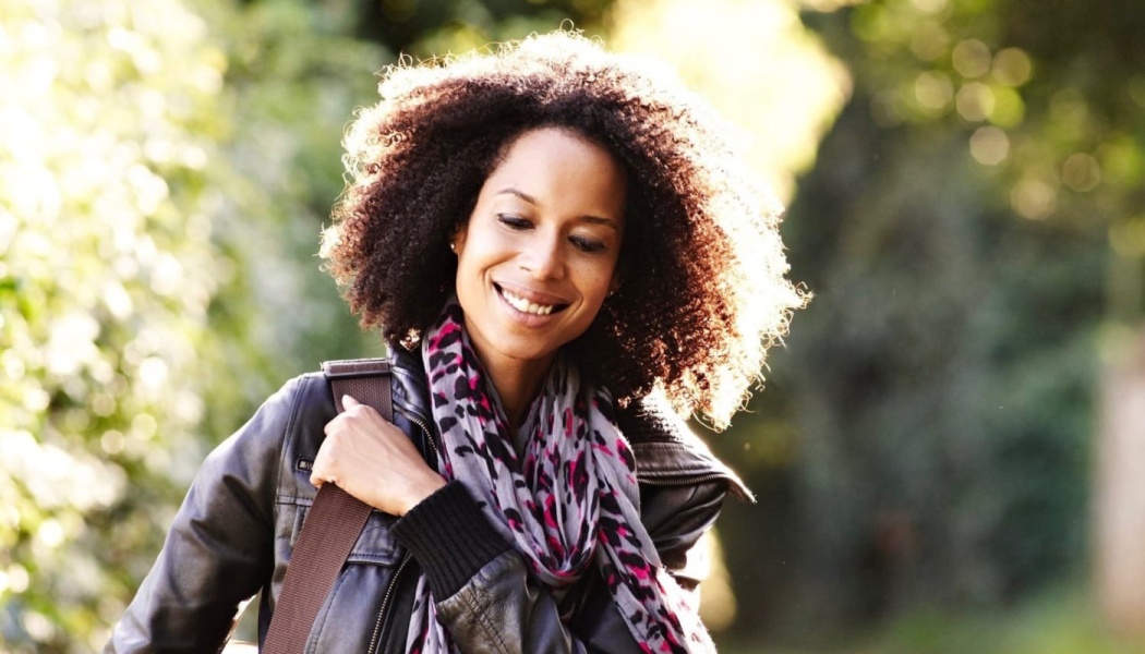 Smiling woman with curly hair wearing a black leather jacket and patterned scarf, walking outdoors in the sunlight.