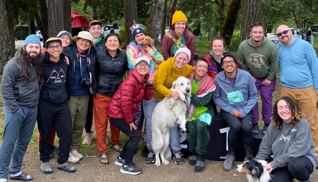A cheerful group of diverse people and their dogs pose together outdoors in a forest setting.
