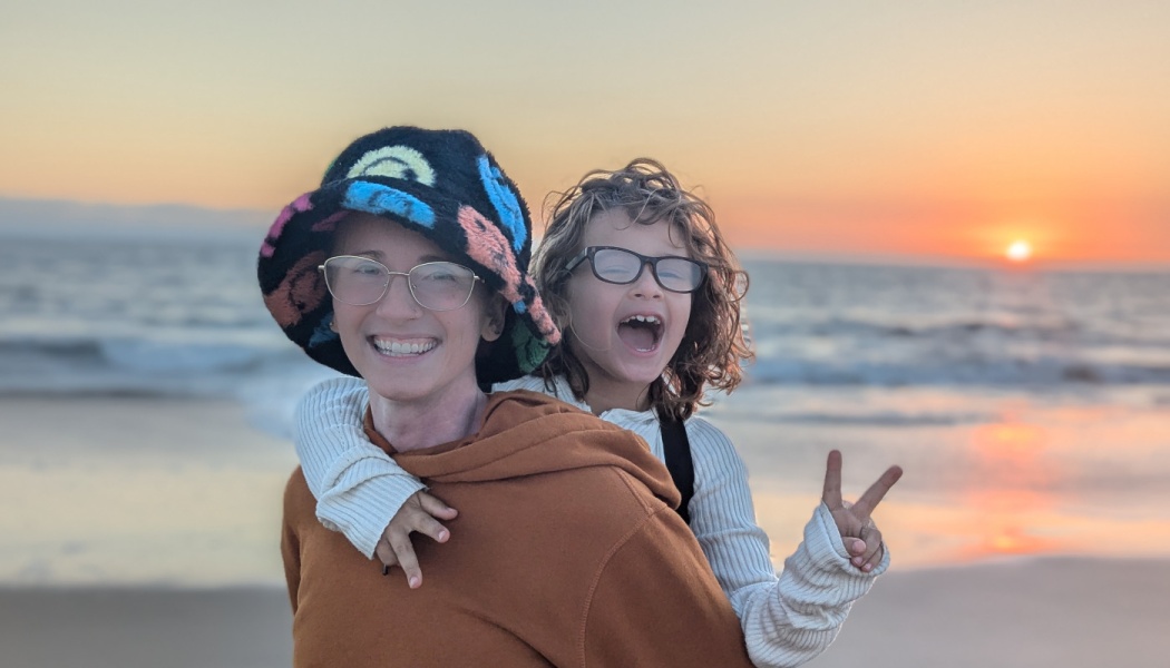 A smiling adult in a colorful bucket hat gives a piggyback ride to a cheering child on a beach at sunset.