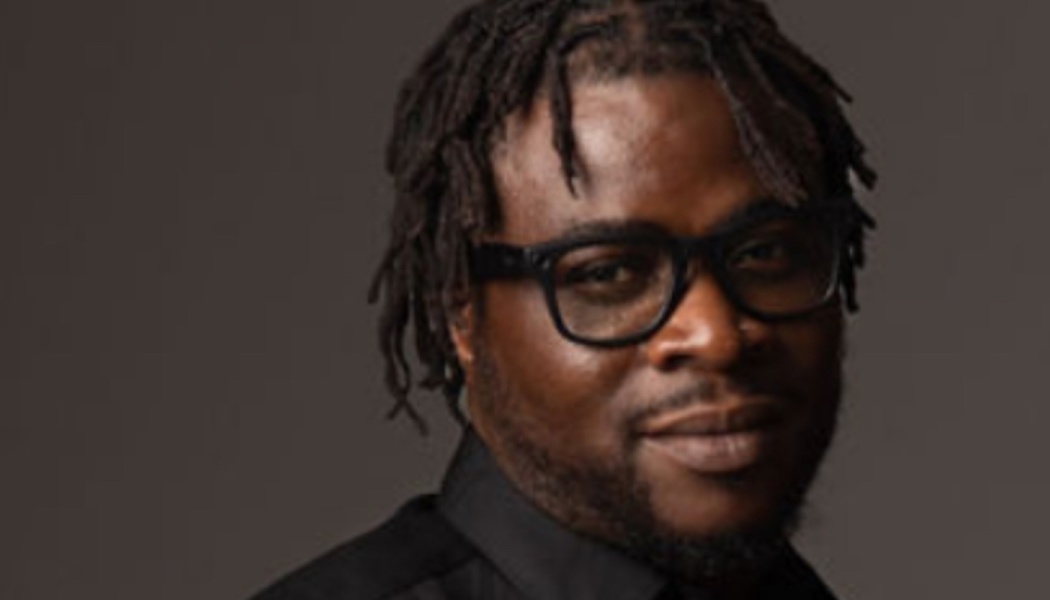 Close-up portrait of a Black man with short dreadlocks and black glasses smiling subtly against a dark background.