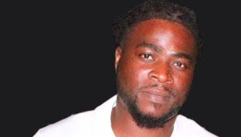 Portrait of a Black man with short dreadlocks and a beard, wearing a white shirt against a black background.