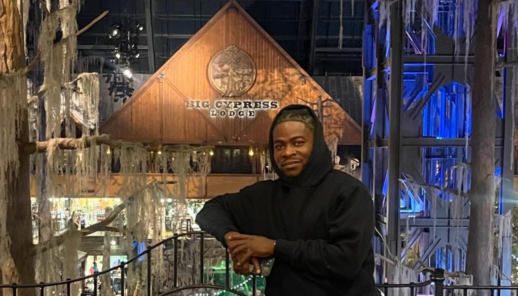 A man in a black hoodie leaning on a balcony railing in front of the wooden Big Cypress Lodge sign.