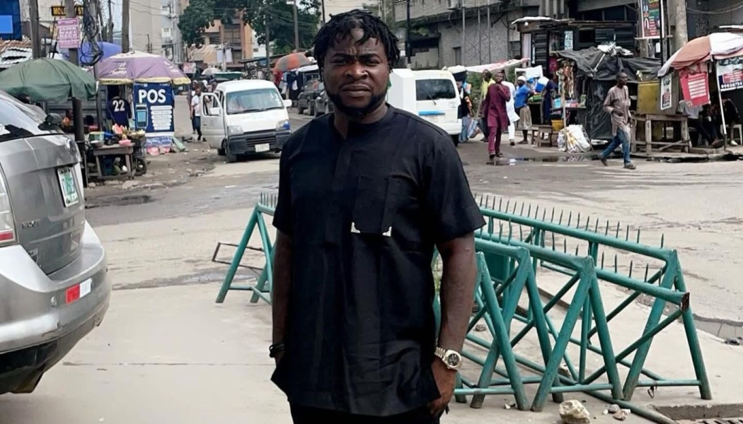 Man with short dreadlocks wearing a black traditional outfit standing on a busy city street with market stalls.