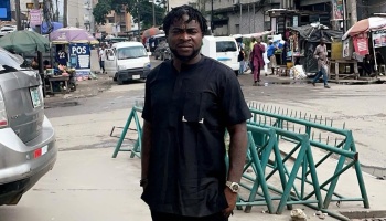 Man with short dreadlocks wearing a black traditional outfit standing on a busy city street with market stalls.