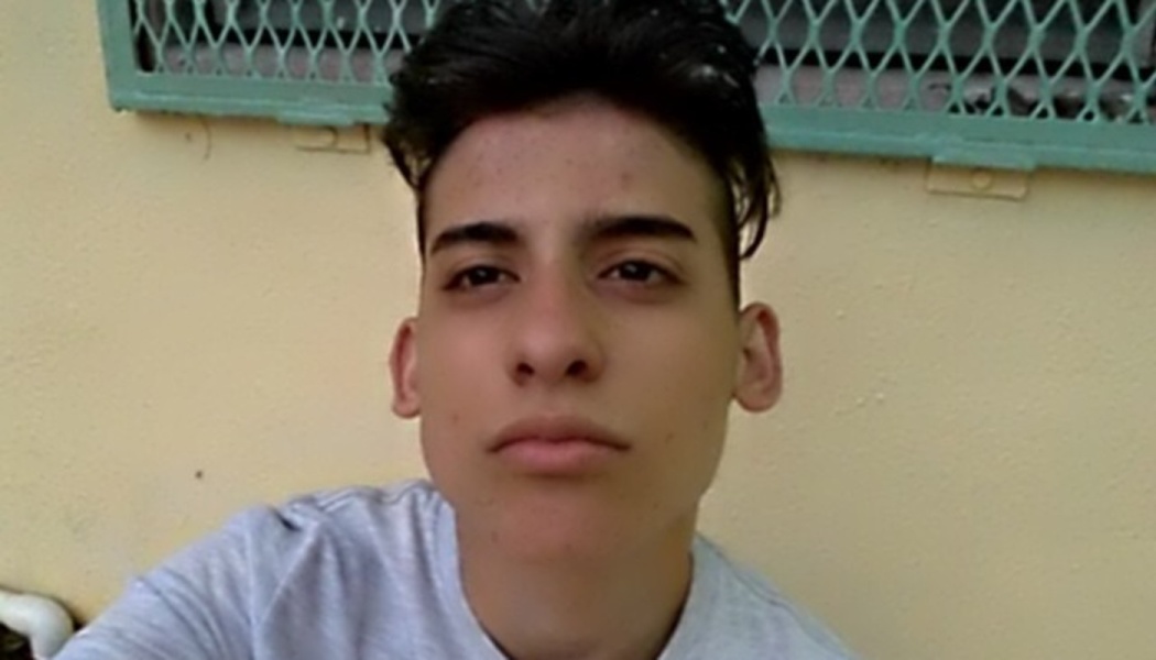 Young man with dark styled hair looking directly at the camera, wearing a light grey t-shirt.