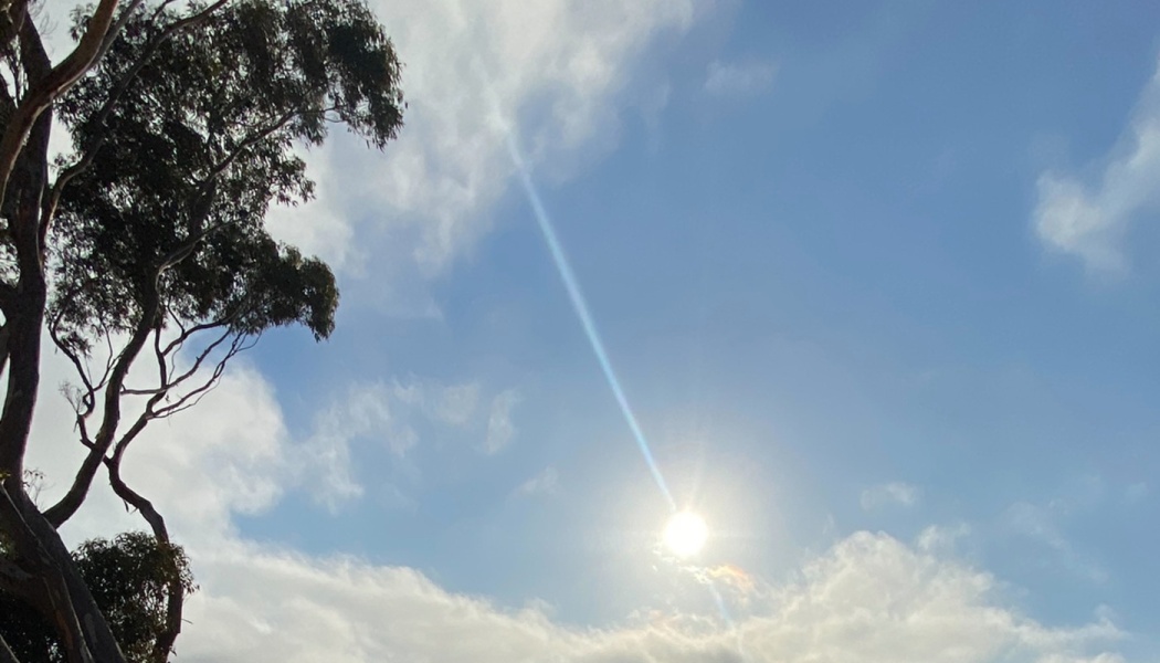 Bright sun emitting a beam of light across a partly cloudy blue sky next to silhouetted tree branches.