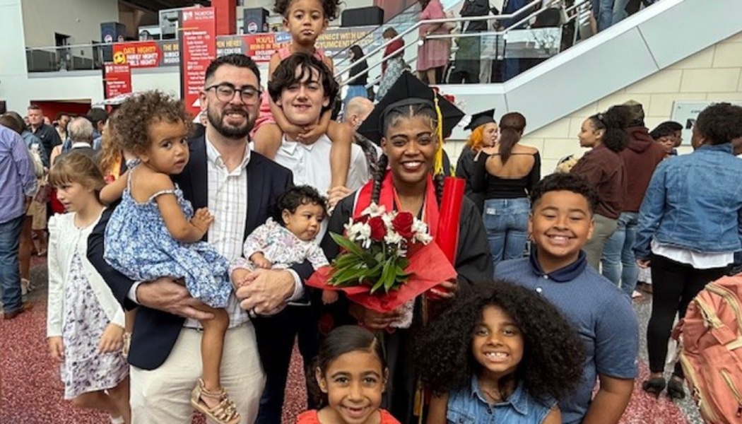 A smiling graduate in a cap and gown holding flowers poses with her large family and young children.