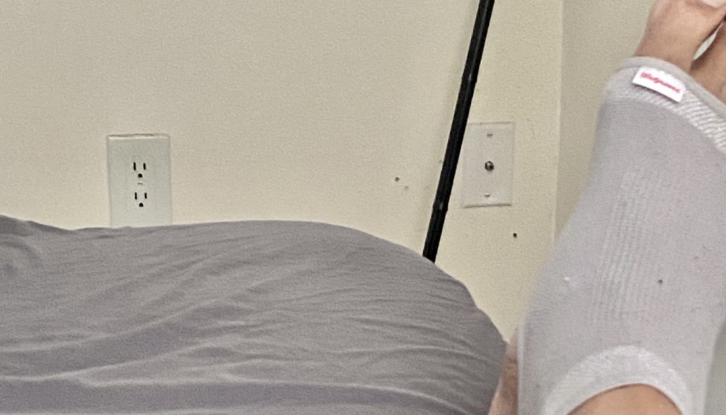 A person's foot wearing a light gray compression ankle sleeve while resting in bed.
