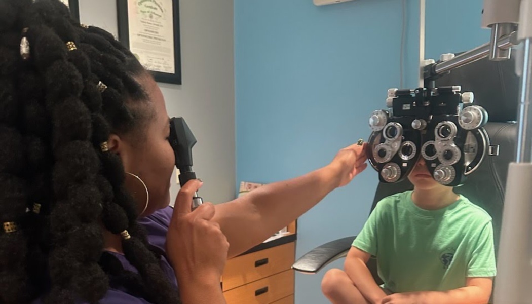 A Black female optometrist conducts a vision exam on a young child using a phoropter.