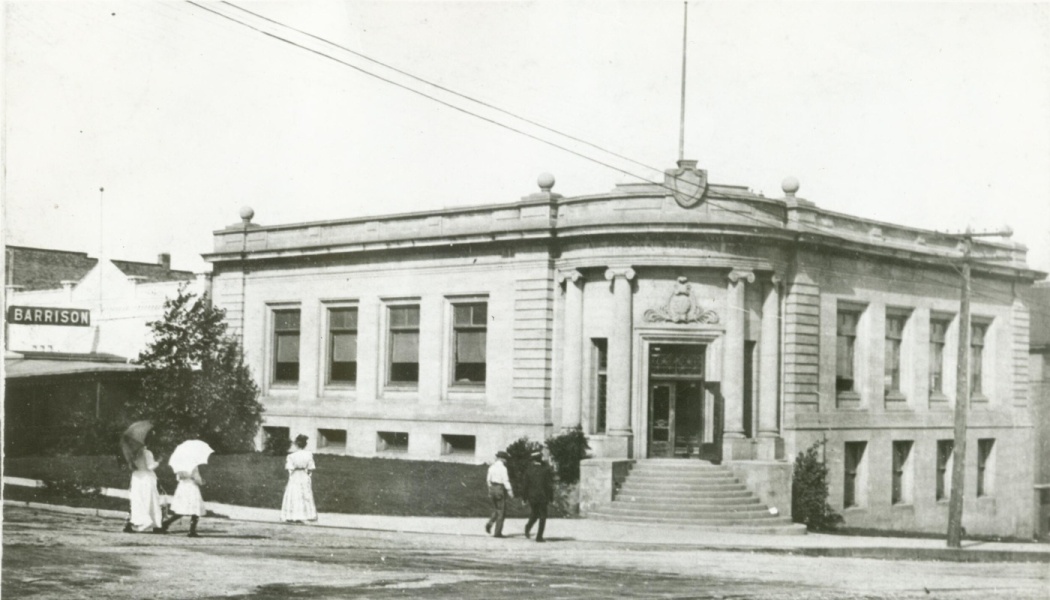 A historic stone building with grand entrance steps and pedestrians in turn-of-the-century attire walking by.