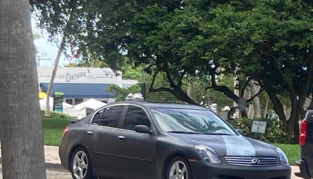 A matte dark grey Infiniti sedan parked on a street with green trees and a building in the background.