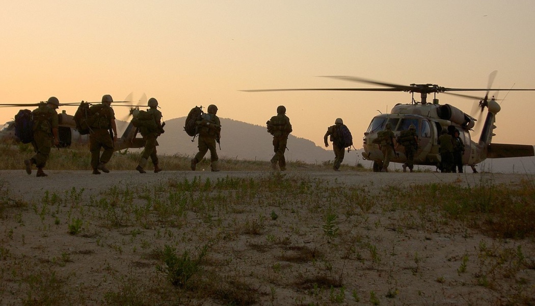 Soldiers in combat gear walk toward military helicopters on a dusty field at sunset.