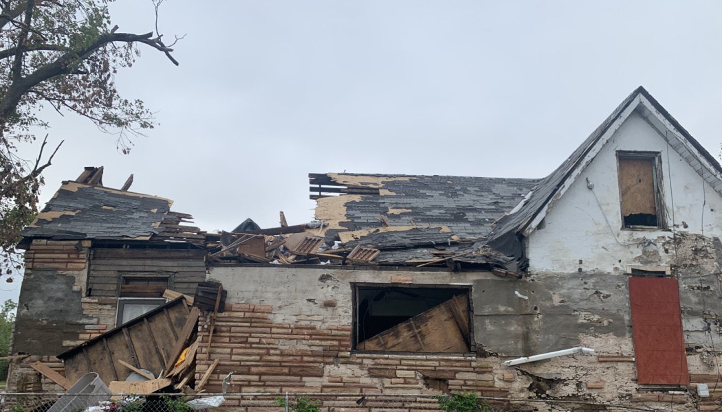 Severely storm-damaged house with a collapsed roof, broken stone siding, and scattered structural debris.