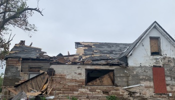Severely storm-damaged house with a collapsed roof, broken stone siding, and scattered structural debris.