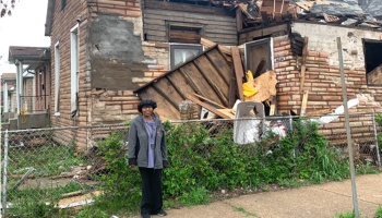 A woman stands in front of a heavily damaged brick house with a collapsed roof and scattered debris.
