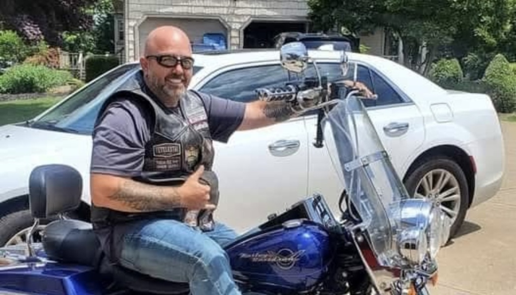 A smiling bald man wearing sunglasses and a leather vest sits on a blue motorcycle in a driveway.