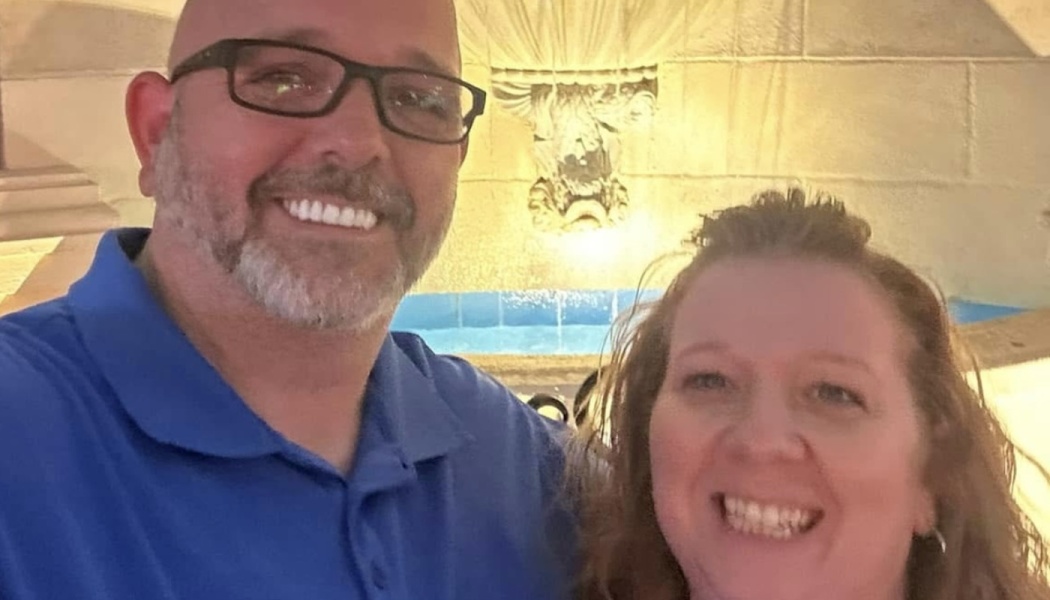A smiling man with glasses in a blue shirt poses for a selfie with a smiling woman in front of a stone fountain.