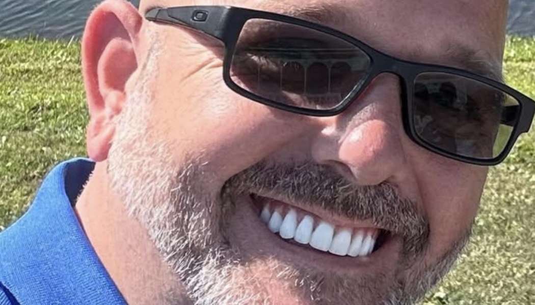 Close-up of a smiling man with a short grey beard and black sunglasses outdoors.