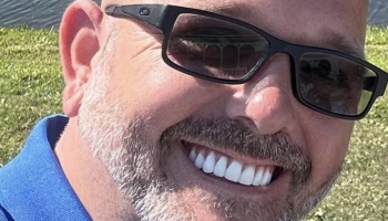 Close-up of a smiling man with a short grey beard and black sunglasses outdoors.
