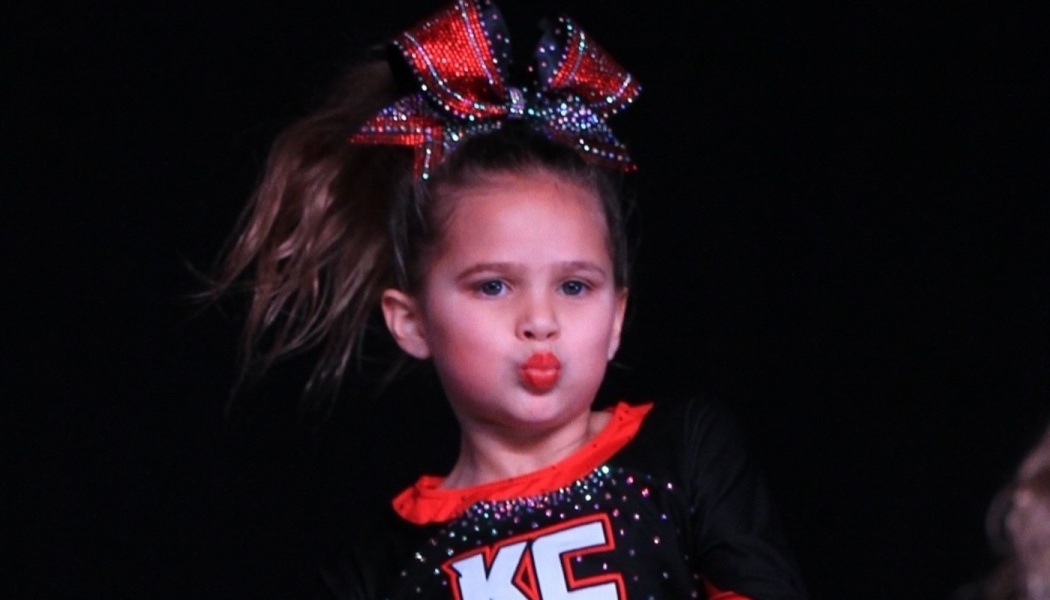 Young cheerleader wearing a large sparkly red bow and cheer uniform making a puckered face.