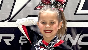 Smiling young cheerleader wearing a red, black, and white uniform with a big sparkly hair bow.