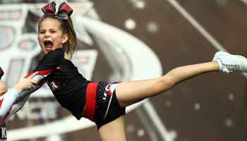 Young cheerleader in a red and black uniform shouting passionately while performing an elevated leg stunt.