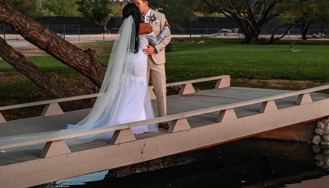 A bride in a white lace gown and long veil kissing her groom in a beige suit on a wooden bridge outdoors.