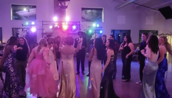 Teenagers in colorful formal dresses and suits dance under purple party lights.