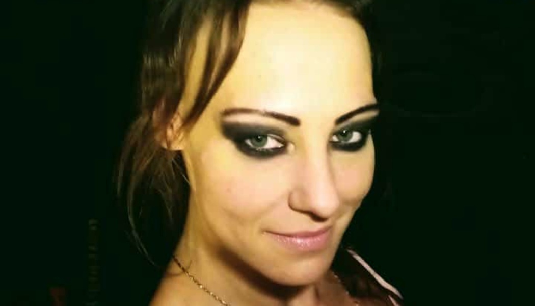 Close-up of a woman with green eyes, dark hair, and smoky eye makeup smiling gently against a dark background.