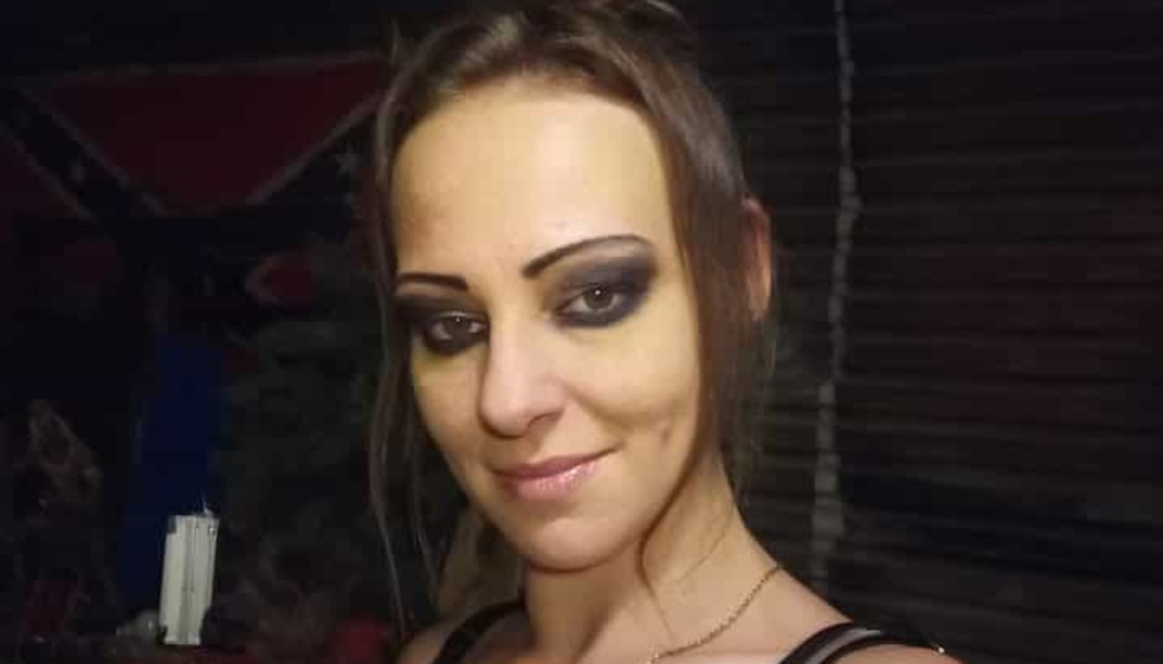 A woman with dark eye makeup smiling at the camera, with a Confederate flag in the background.