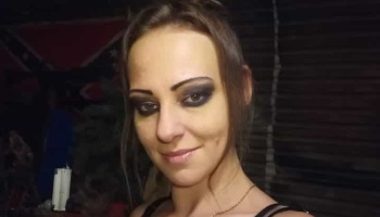 A woman with dark eye makeup smiling at the camera, with a Confederate flag in the background.