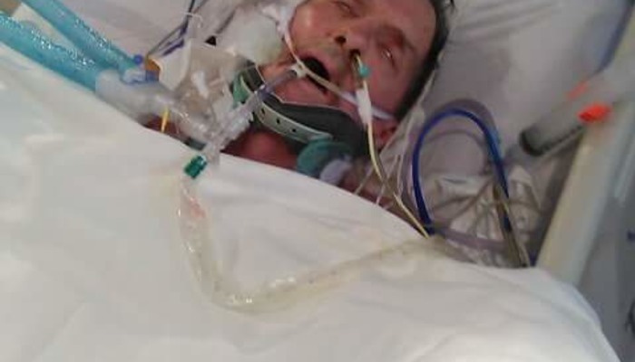 A man lies in a hospital bed wearing a neck brace and connected to breathing tubes.
