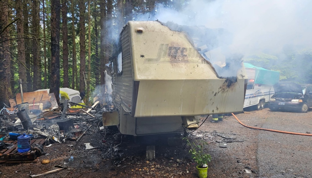 A heavily fire-damaged RV trailer billows smoke surrounded by charred debris in a forest campsite.