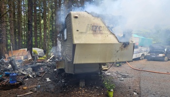 A heavily fire-damaged RV trailer billows smoke surrounded by charred debris in a forest campsite.