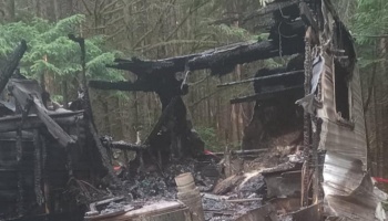 Charred, burnt remains of a trailer home completely destroyed by fire in a wooded area.