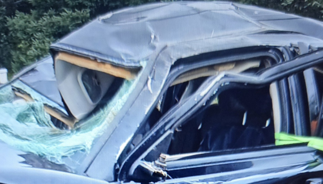 Heavily damaged dark car with a crushed roof and shattered windshield after a severe crash.