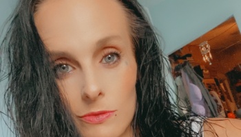 Close-up portrait of a woman with wet dark hair, light eyes, and pink lipstick looking at the camera.