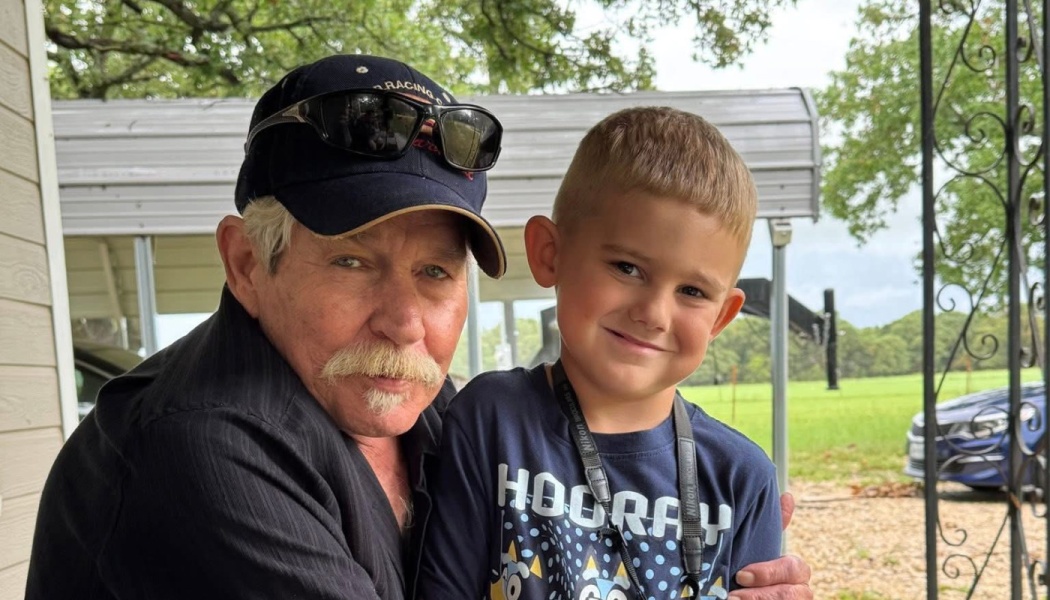 An older man with a mustache and cap hugs a smiling young boy outdoors.