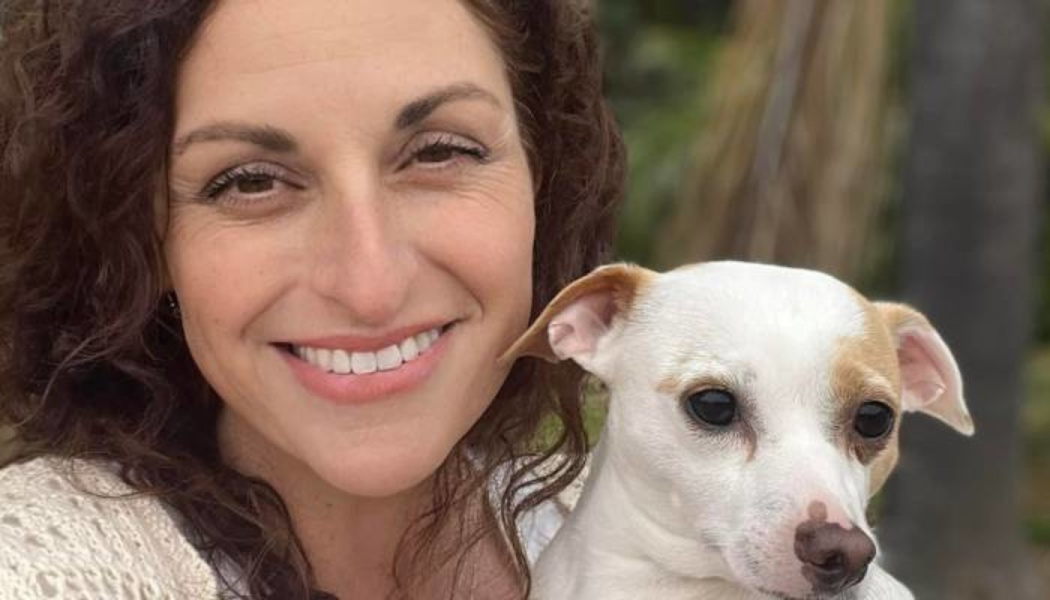 A smiling woman with dark curly hair poses closely with a small white and tan dog outdoors.
